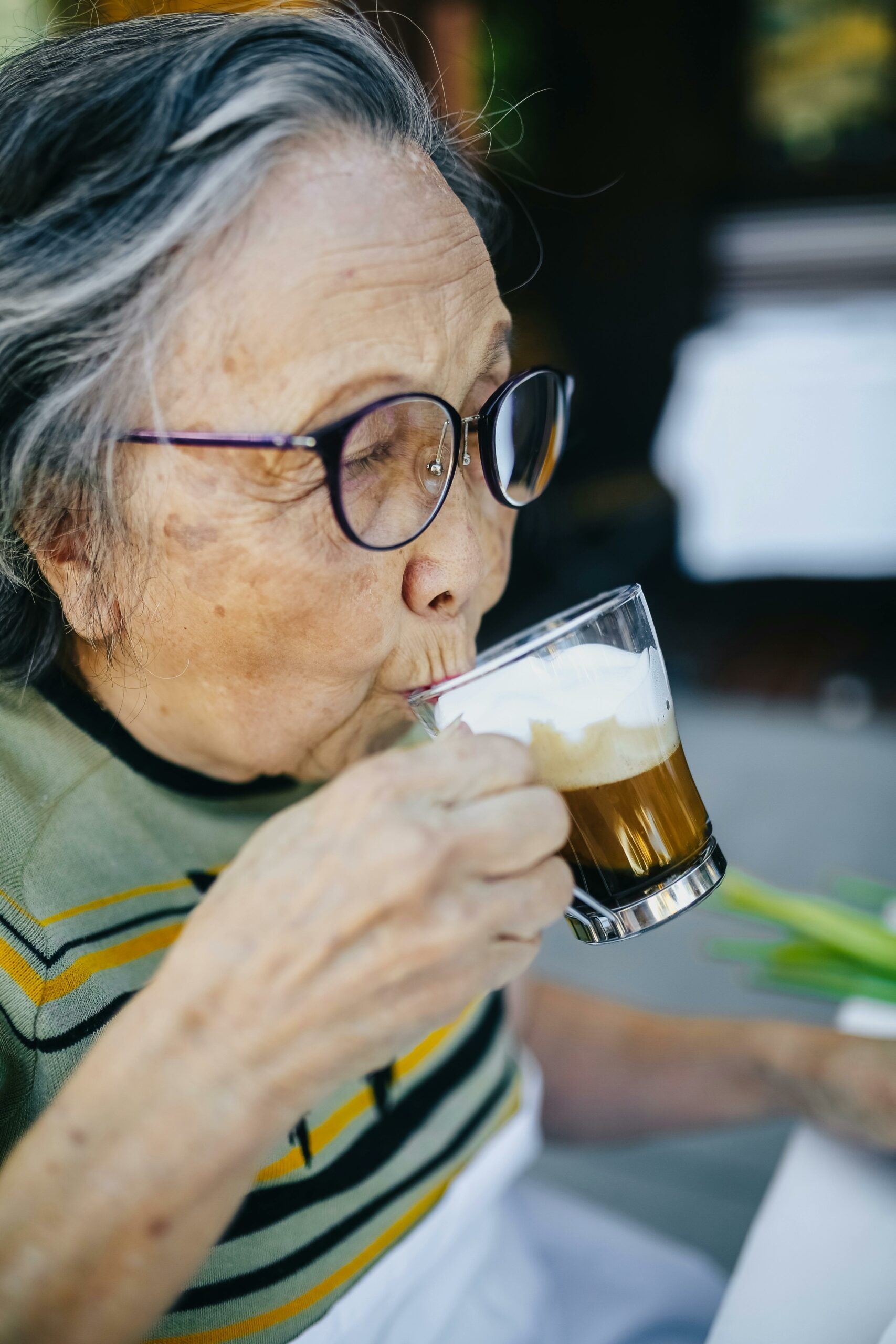 Senior Asian woman sipping cappuccino outdoors, enjoying leisure time at a cafe.